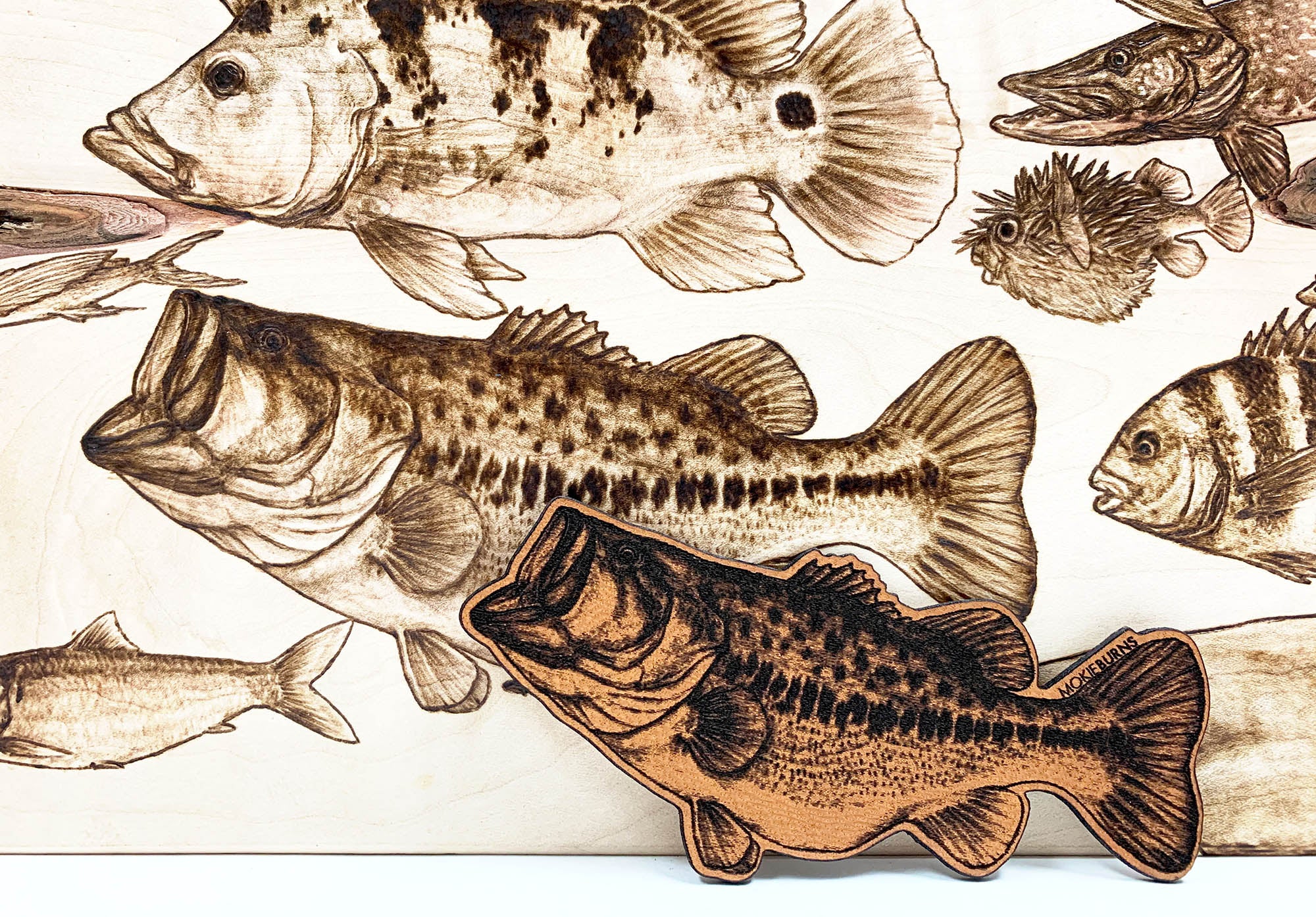 a photo showcasing a unique and detailed genuine leather patch of a largemouth bass fish, with the original wood burning art behind it. This patch is used for fishing hats by Mokie Burns™