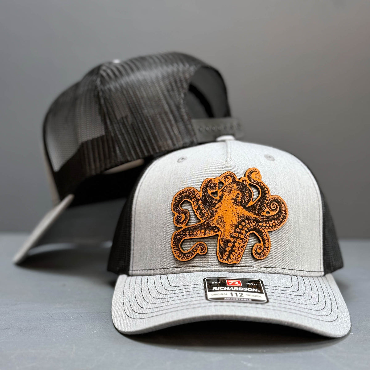 Close-up image of a Richardson 112 trucker hat in heather grey and black, featuring a striking leather patch with an octopus design, also known as &#39;kraken&#39;, perfect for recreational saltwater fishermen seeking a unique and marine-themed cap for their fishing outings.