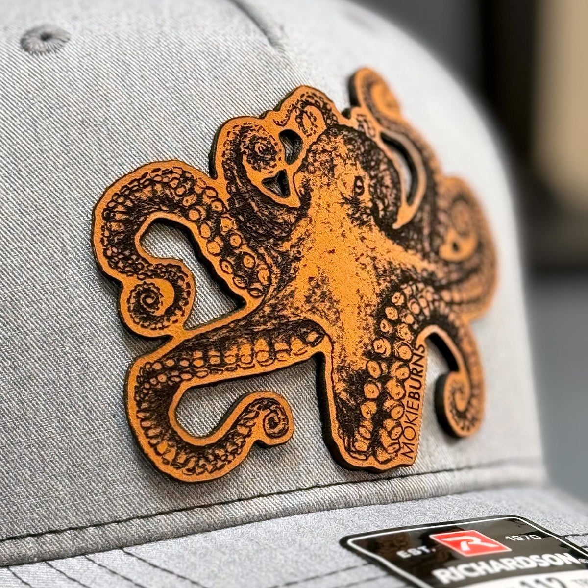 Close-up image of a Richardson 112 trucker hat in heather grey and black, featuring a striking leather patch with an octopus design, also known as &#39;kraken&#39;, perfect for recreational saltwater fishermen seeking a unique and marine-themed cap for their fishing outings.
