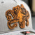 Close-up image of a Richardson 112 trucker hat in heather grey and black, featuring a striking leather patch with an octopus design, also known as 'kraken', perfect for recreational saltwater fishermen seeking a unique and marine-themed cap for their fishing outings.