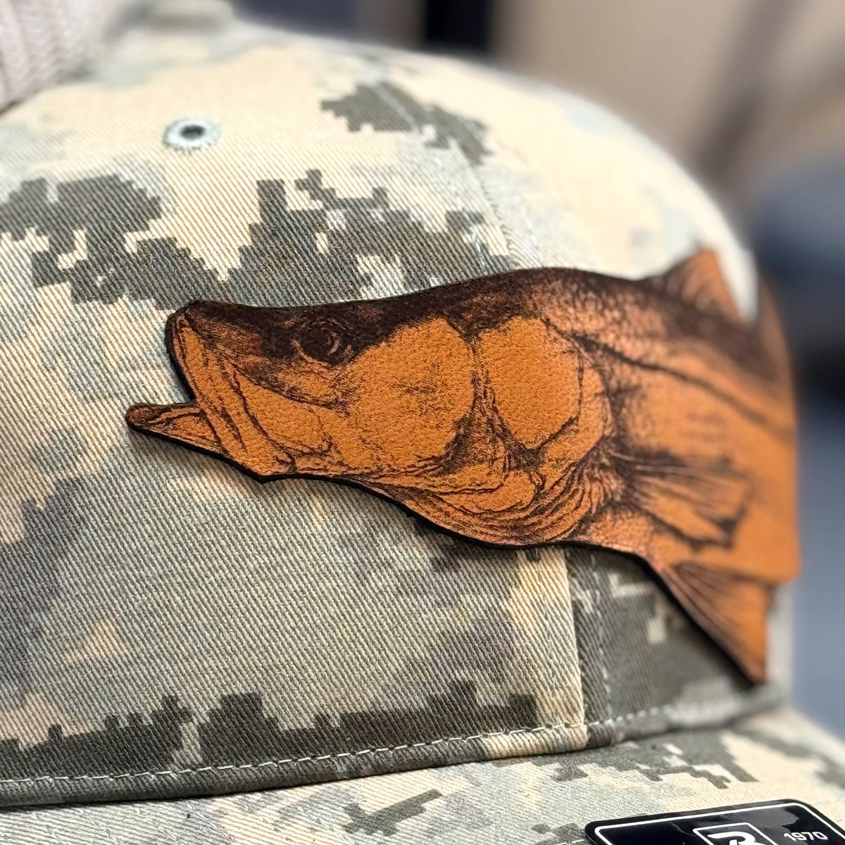 Photo of a Richardson 112 trucker hat in digital khaki camo colorway featuring a detailed leather patch of a snook, ideal for recreational saltwater fishermen seeking themed and functional headwear for their fishing trips.